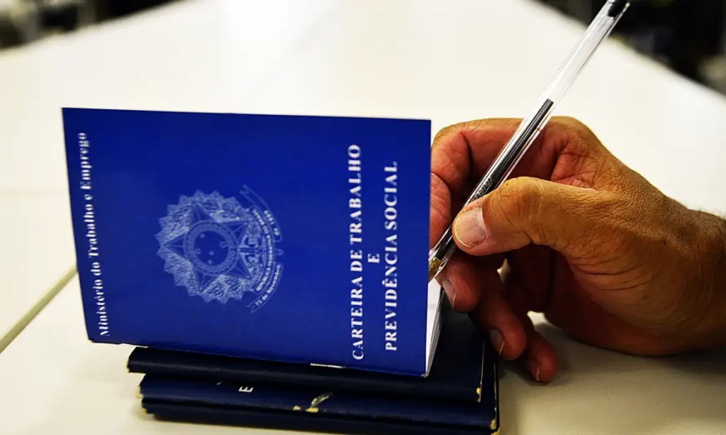 A imagem mostra uma pessoa com uma caneta assinando a na mão carteira de trabalho, a chamada CLT.