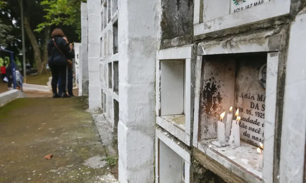 A foto mostra pessoas visitando túmulos em cemitério municipal de São Paulo. Em um dos túmulos, há velas acesas.