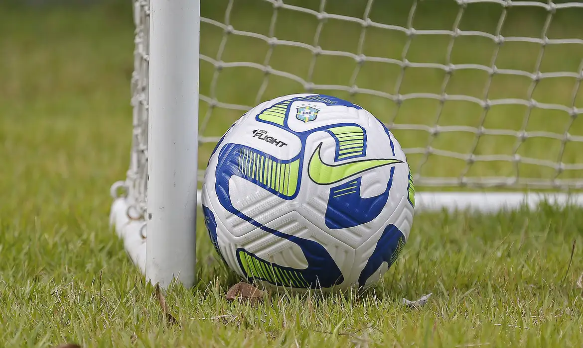 A foto mostra uma bola de futebol branca com detalhes azul e verde junto à uma trave branca.