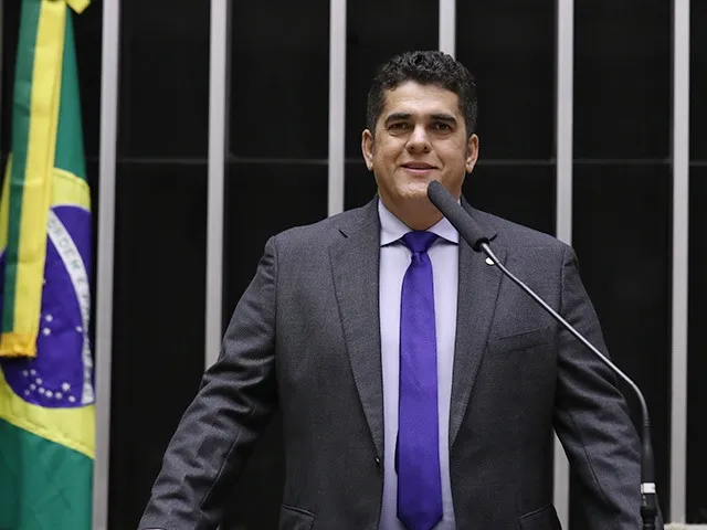 homem branco, de pé discursando da tribuna da Câmara dos Deputados, veste terno cinza, camisa branca e gravata azul