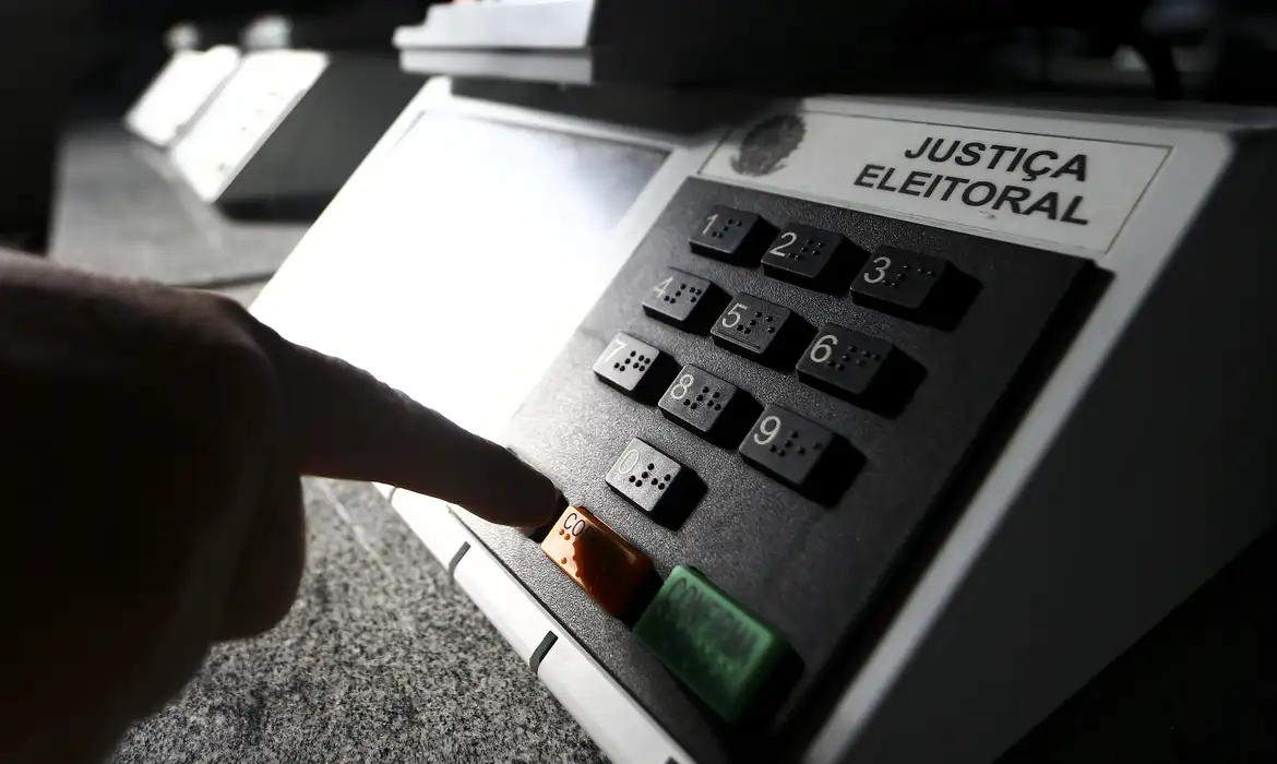 A foto mostra a mão de uma pessoa usando uma urna eletrônica.