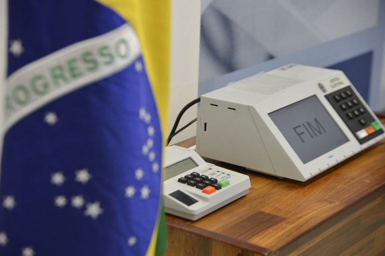 Urna eletrônica sobre uma mesa ao lado da bandeira do Brasil